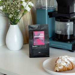 A Dimattina Single Origin coffee from Kenya in a kitchen setting, with a white vase of flowers and a mocca master